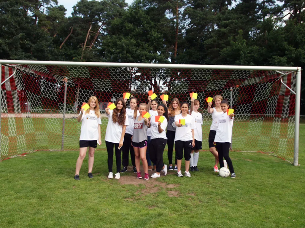 Schülergruppe in weißen T-Shirts steht mit erhobenen Armen vor einem Fußballtor.